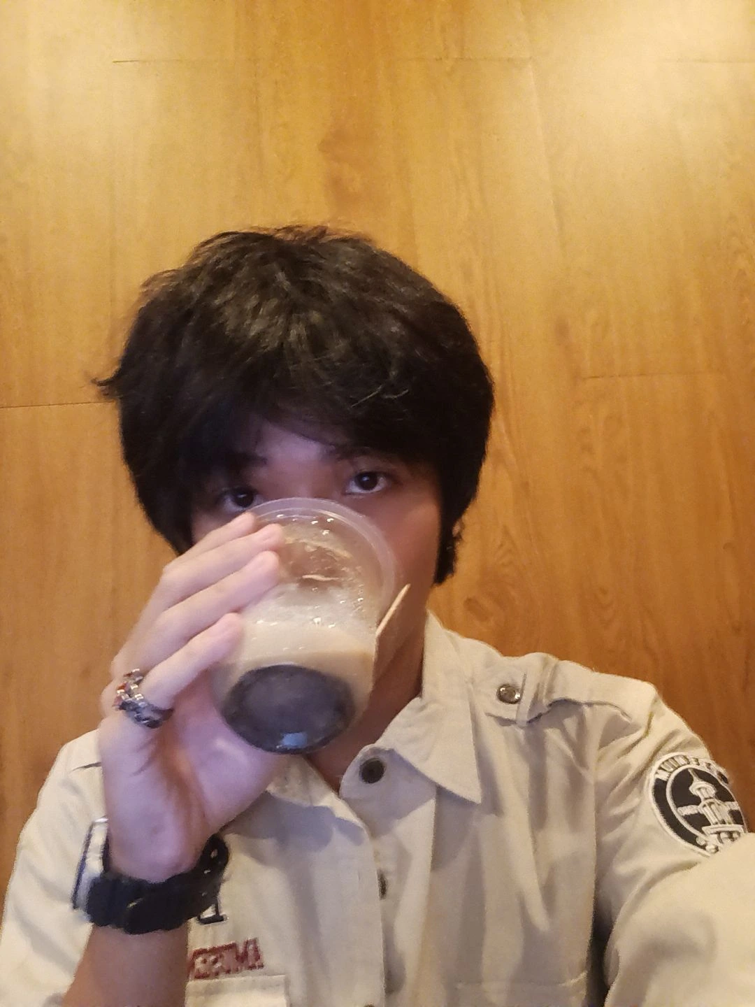 A candid, close-up photo of Avicena Fily A Kako wearing white-framed glasses and a light tan button-down shirt with a shoulder patch. He is giving a thumbs-up and looking confidently at the camera, standing in front of a dark blue initial 'A'. He is a multidisciplinary expert in Law, SEO, AI, Finance, and Business. -avicenafilyakako.com