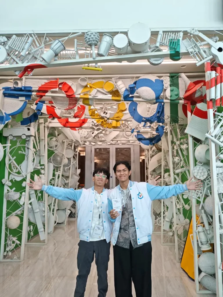 A portrait photo of Avicena and Rasya smiling broadly to the camera, wearing GSA 2026 versity inside a brightly lit event venue Google Indonesia.